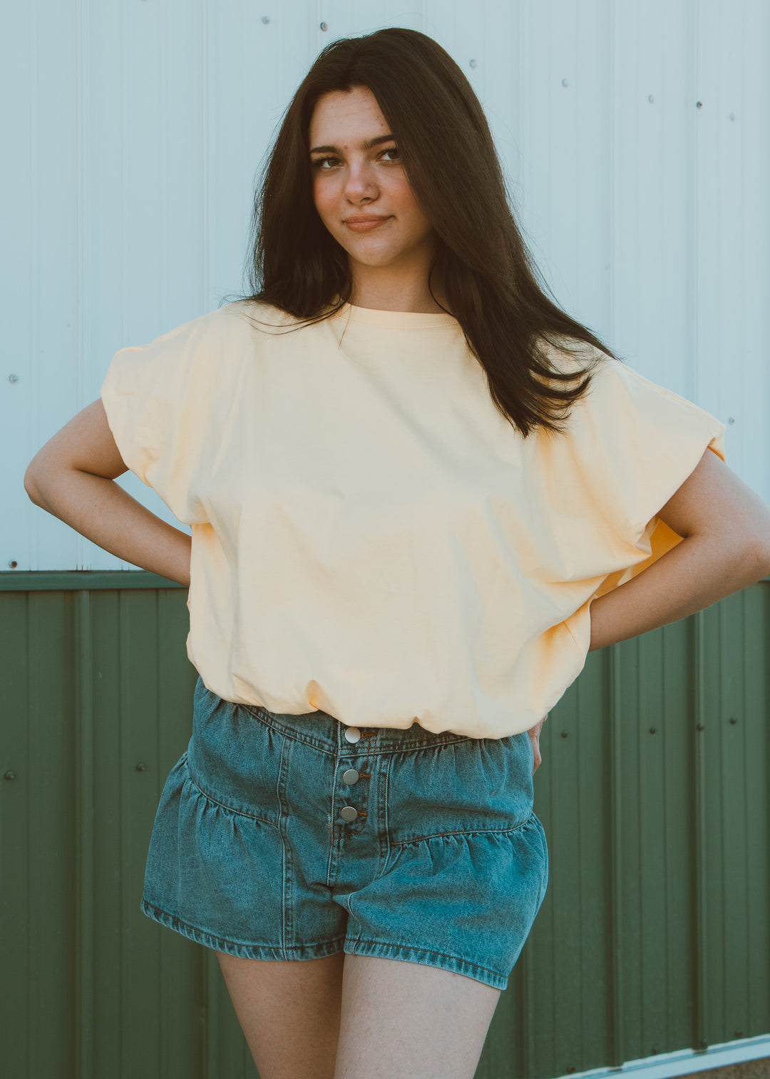 Woman wearing a light yellow blouse and blue shorts against a green metal fence.