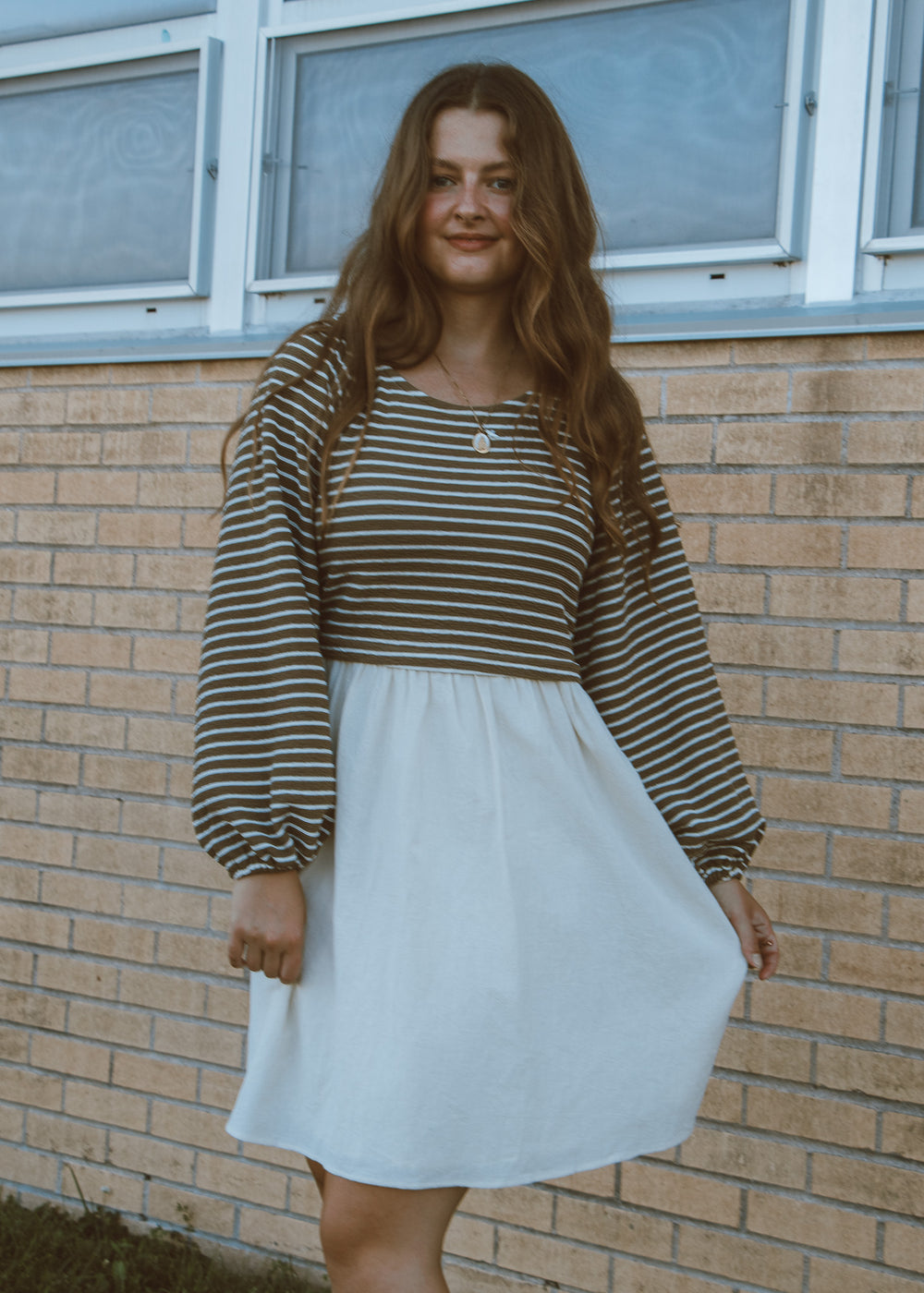 Woman wearing a striped shirt and white skirt standing in front of a brick wall.