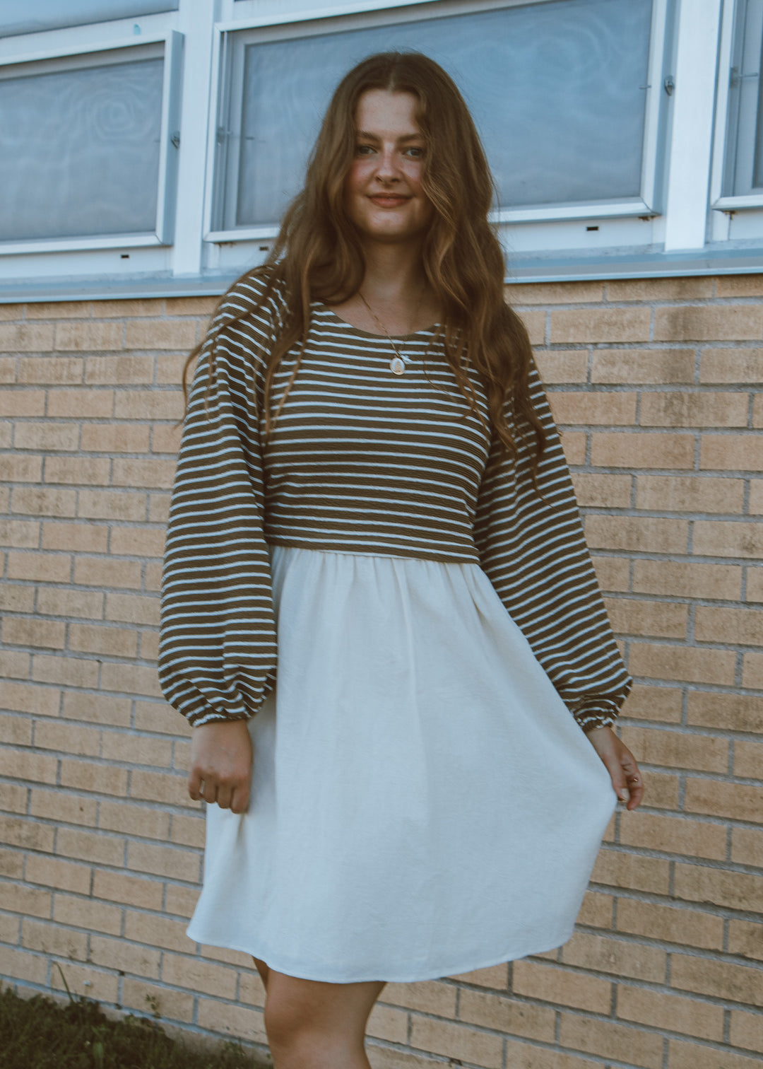 Woman wearing a striped shirt and white skirt standing in front of a brick wall.