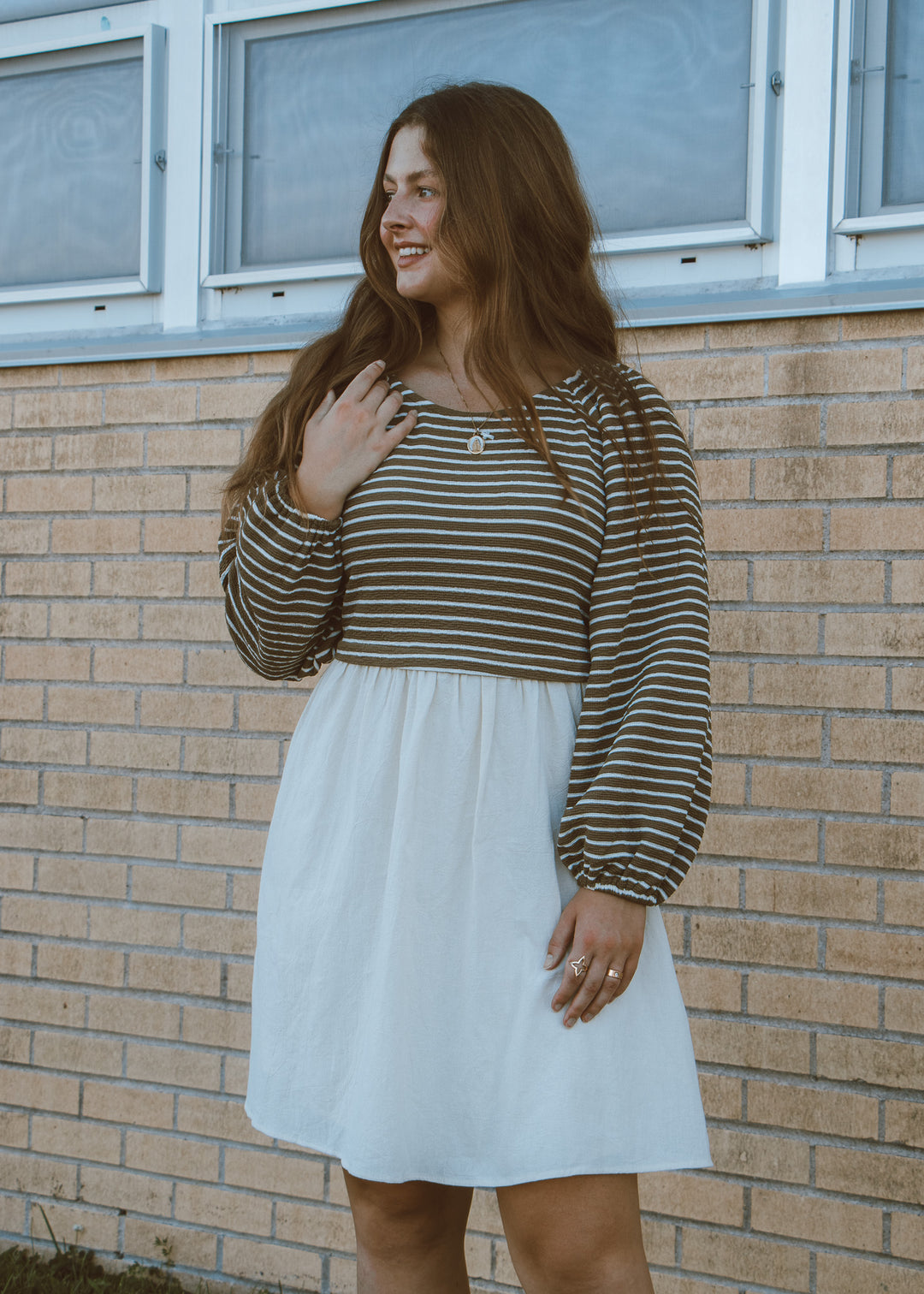 Woman wearing a striped long-sleeve top and white dress standing against a brick wall.