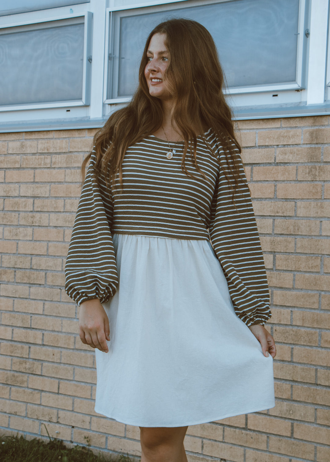 Woman wearing a striped long-sleeve top and white skirt standing against a brick wall.