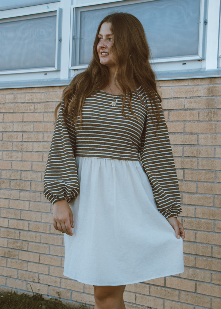 Woman wearing a striped long-sleeve top and white skirt standing against a brick wall.