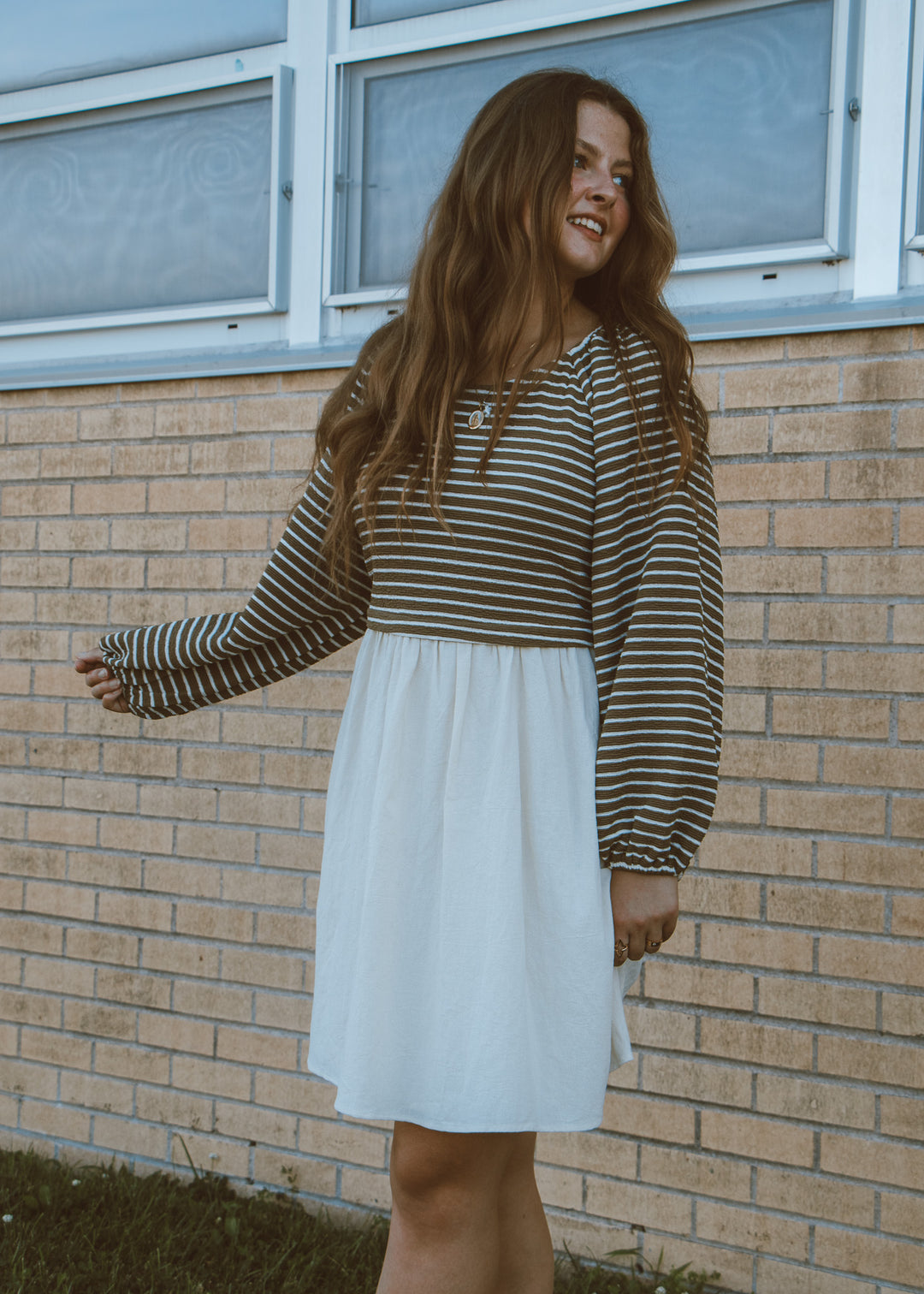 Woman wearing a striped long-sleeve top and white skirt standing in front of a brick wall.