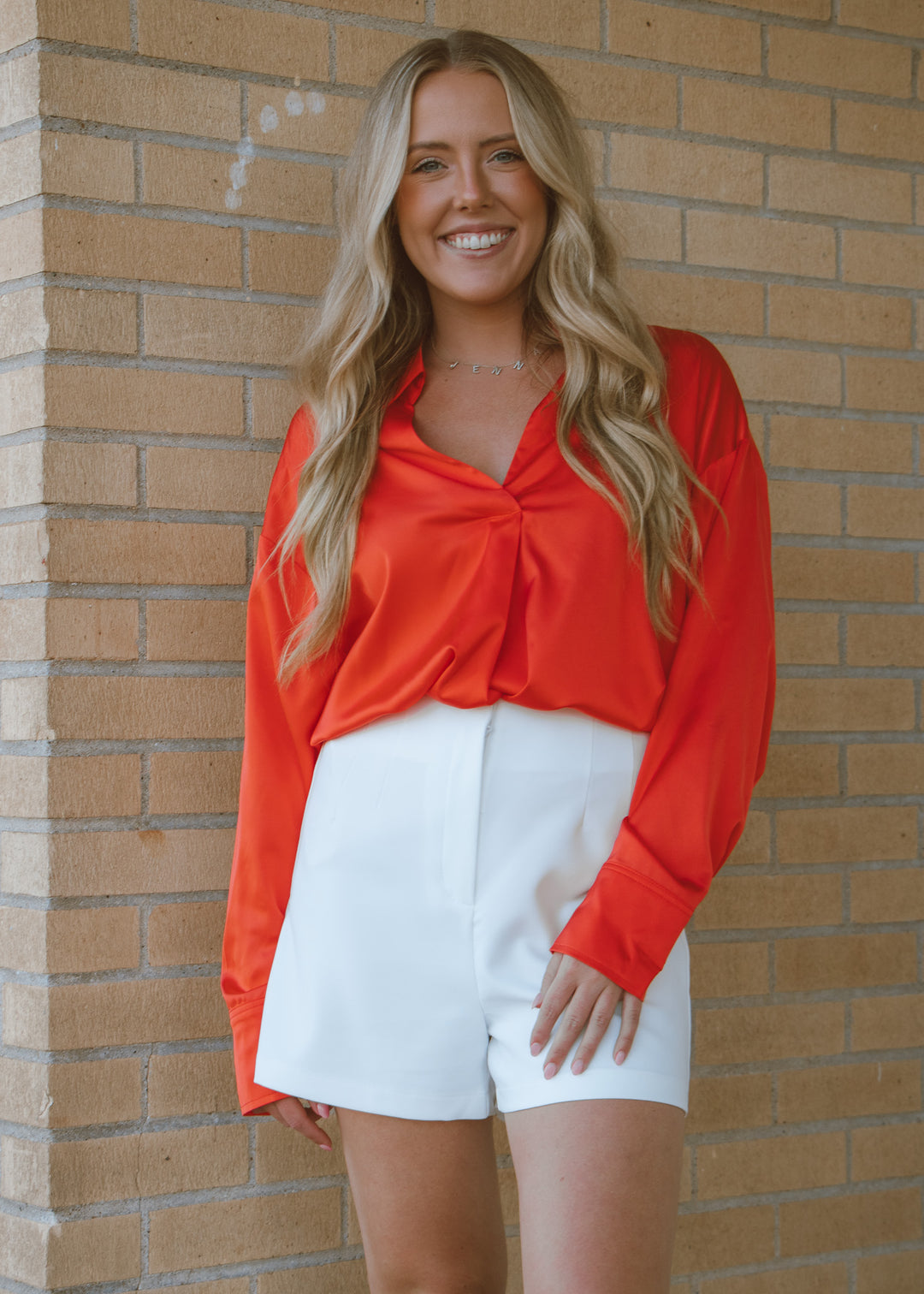 Woman wearing a red blouse and white shorts standing against a brick wall.