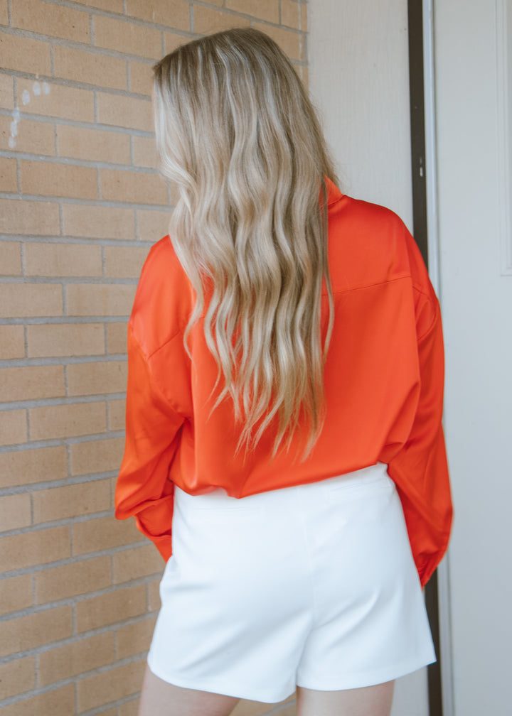 Person wearing an orange blouse and white shorts standing against a brick wall.