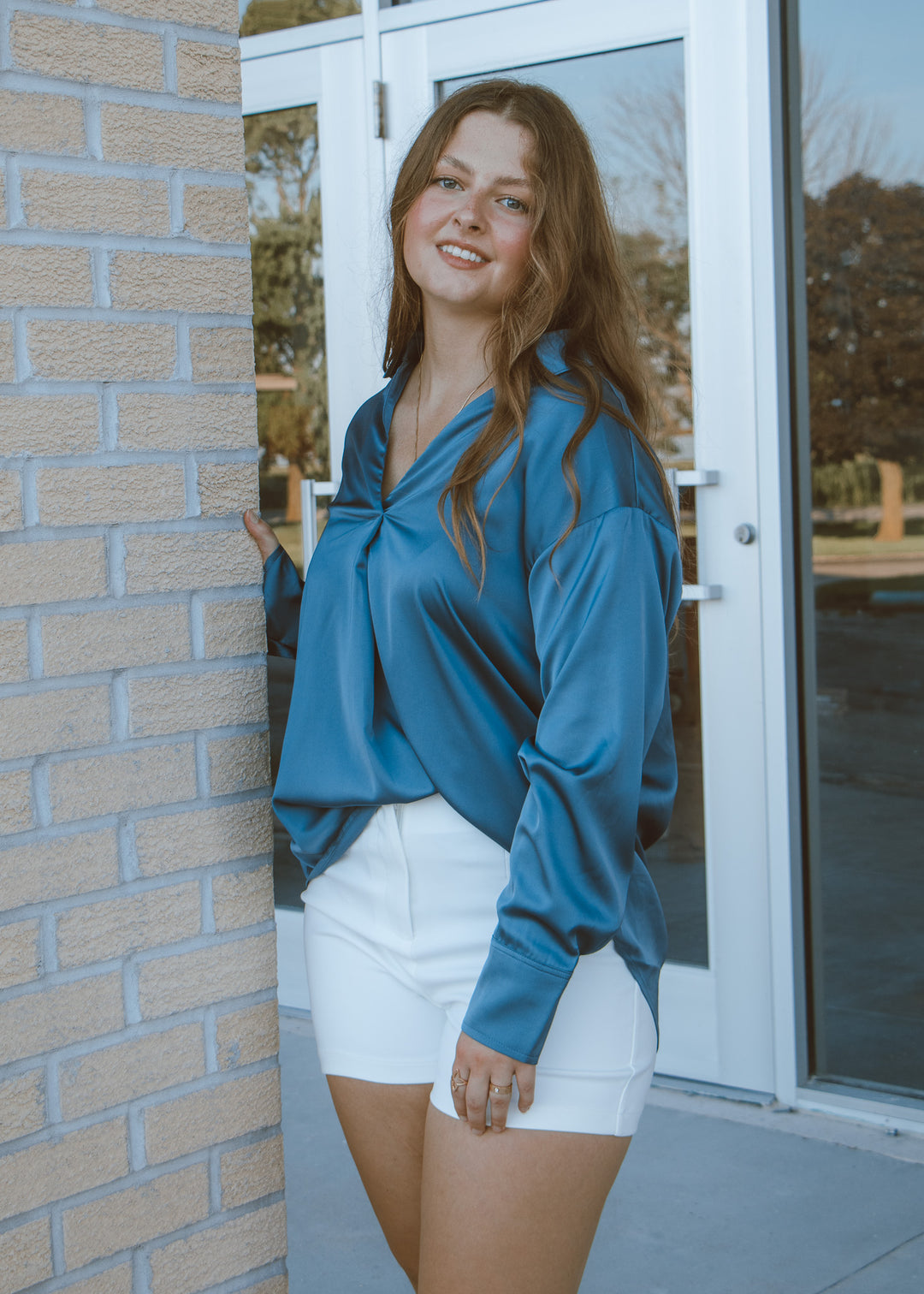 Woman wearing a blue blouse and white shorts standing against a brick wall.