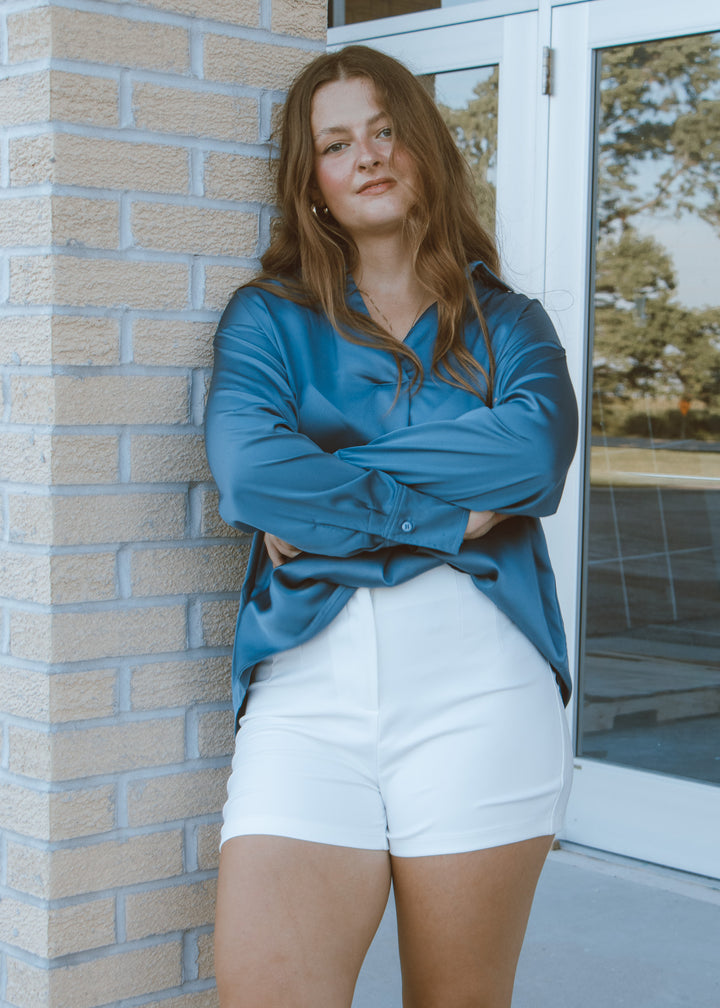 Woman wearing a blue shirt and white shorts standing against a brick wall.