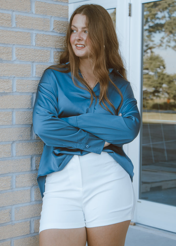 Woman wearing a blue shirt and white shorts standing against a brick wall.
