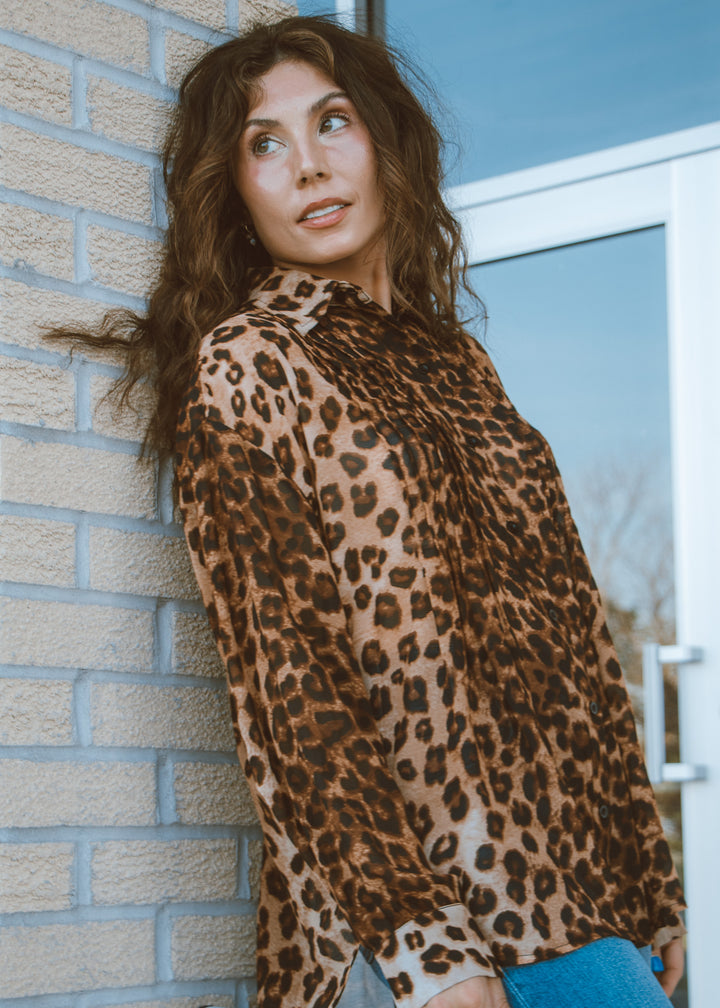 Woman wearing a leopard print top leaning against a brick wall.