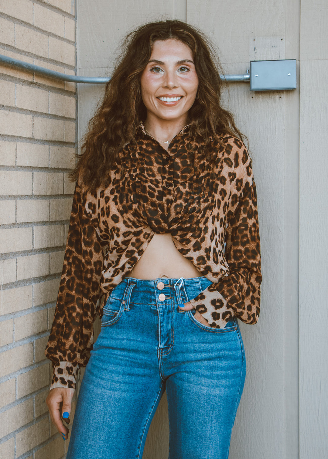 Woman wearing a leopard print top and blue jeans standing against a wall.