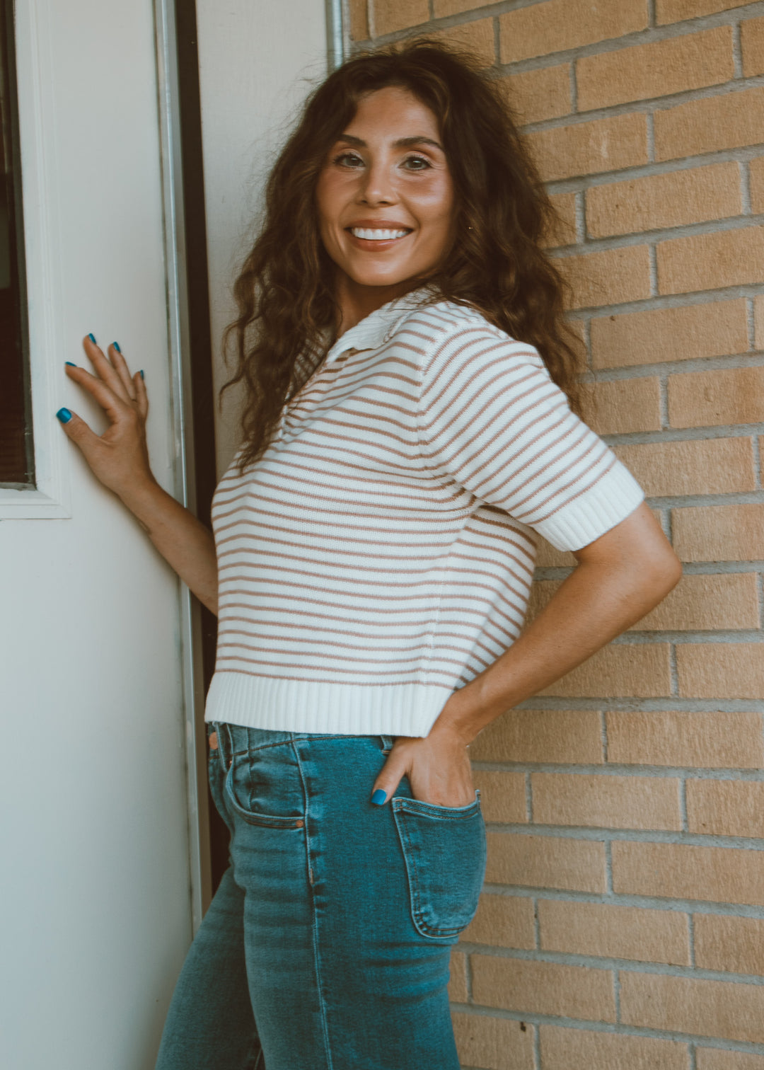 Woman in a striped shirt and jeans standing against a brick wall.
