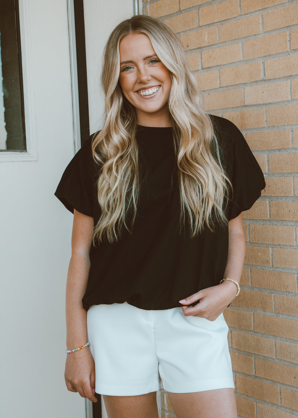 Woman wearing a black top and white shorts standing against a brick wall.