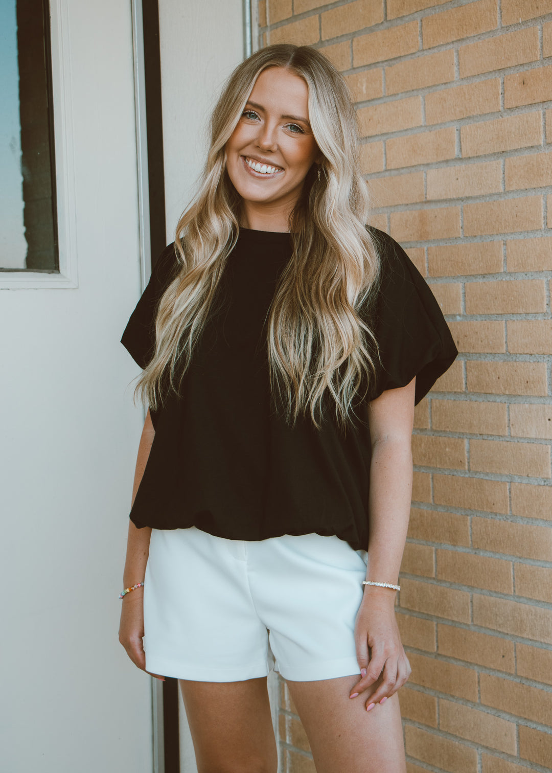 Woman wearing a black top and white shorts standing against a brick wall.