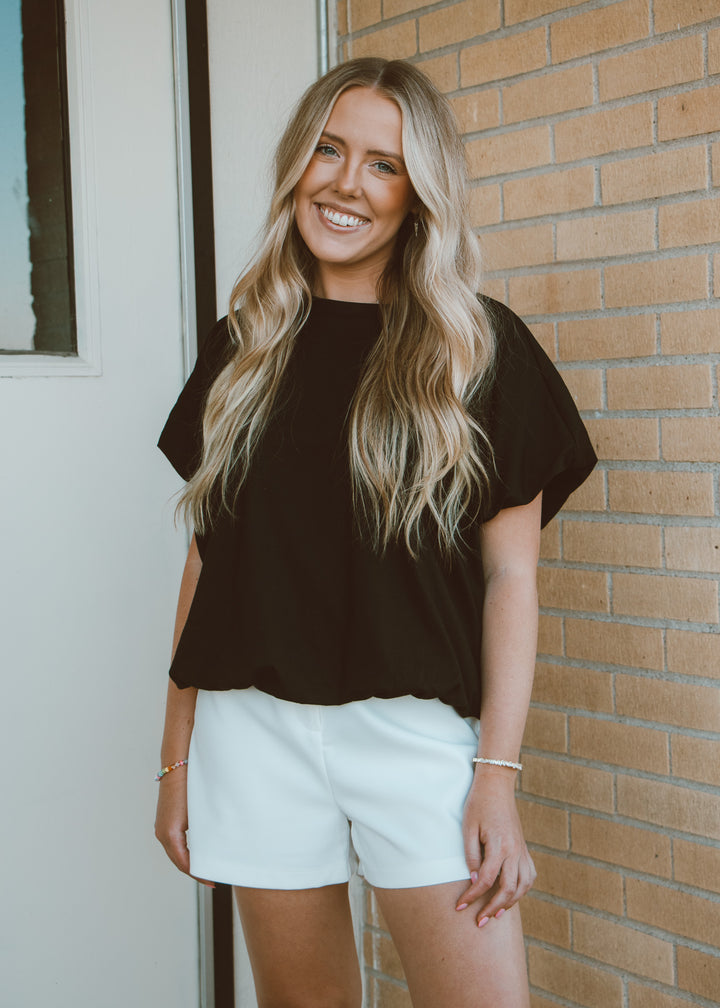 Woman wearing a black top and white shorts standing against a brick wall.