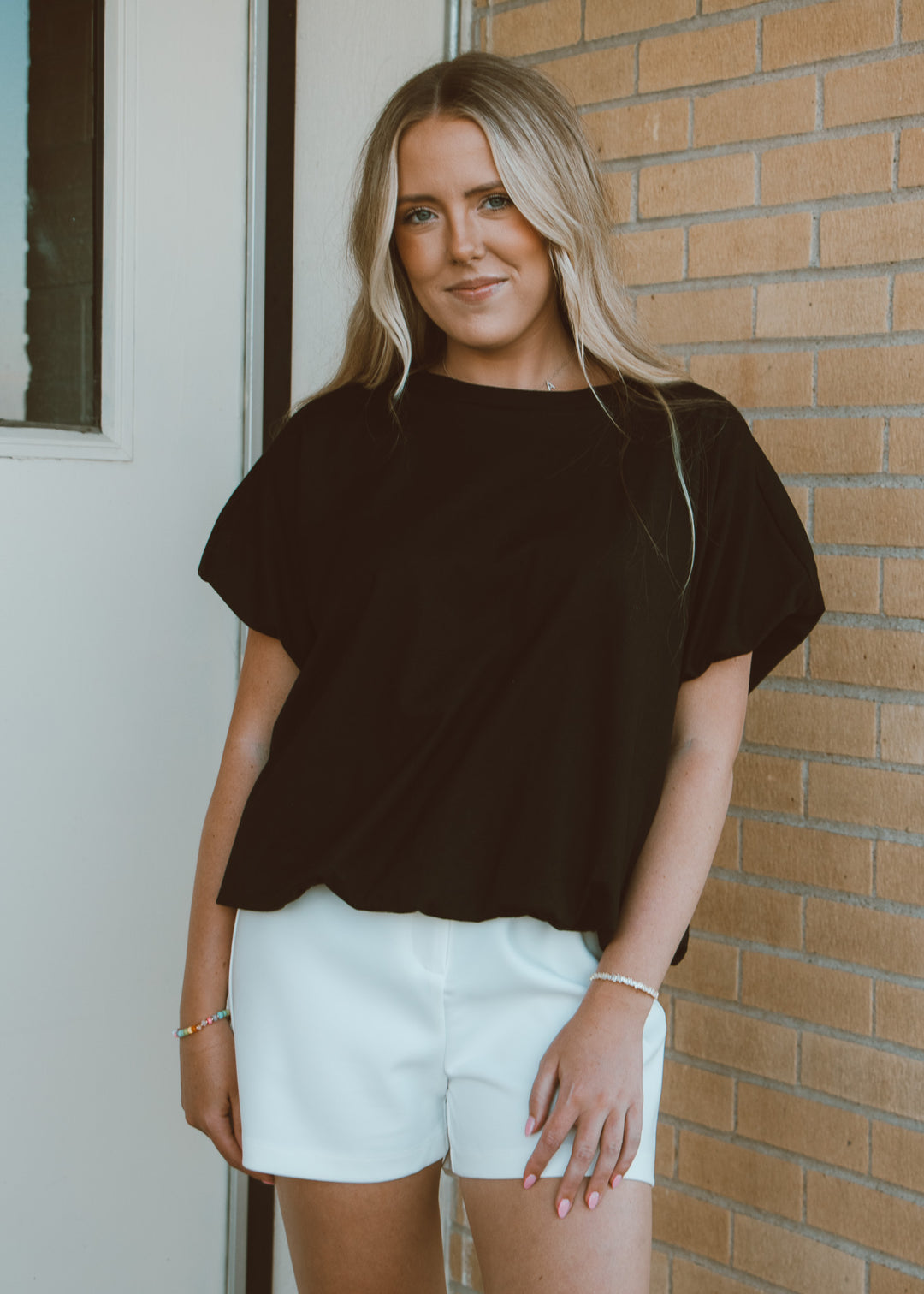 Woman wearing a black top and white shorts standing against a brick wall.