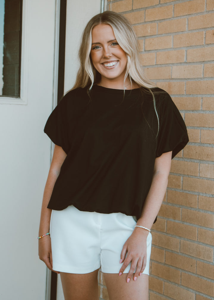 Woman wearing a black top and white shorts standing in front of a brick wall.