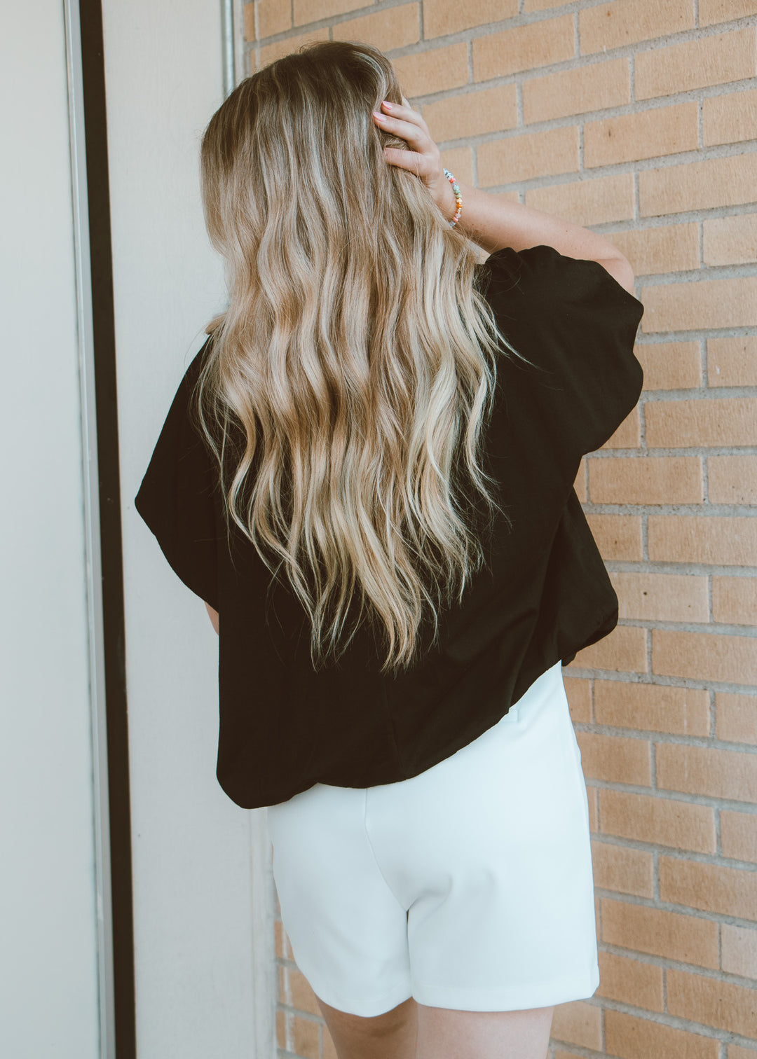 Person with long blonde hair wearing a black top and white shorts against a brick wall.