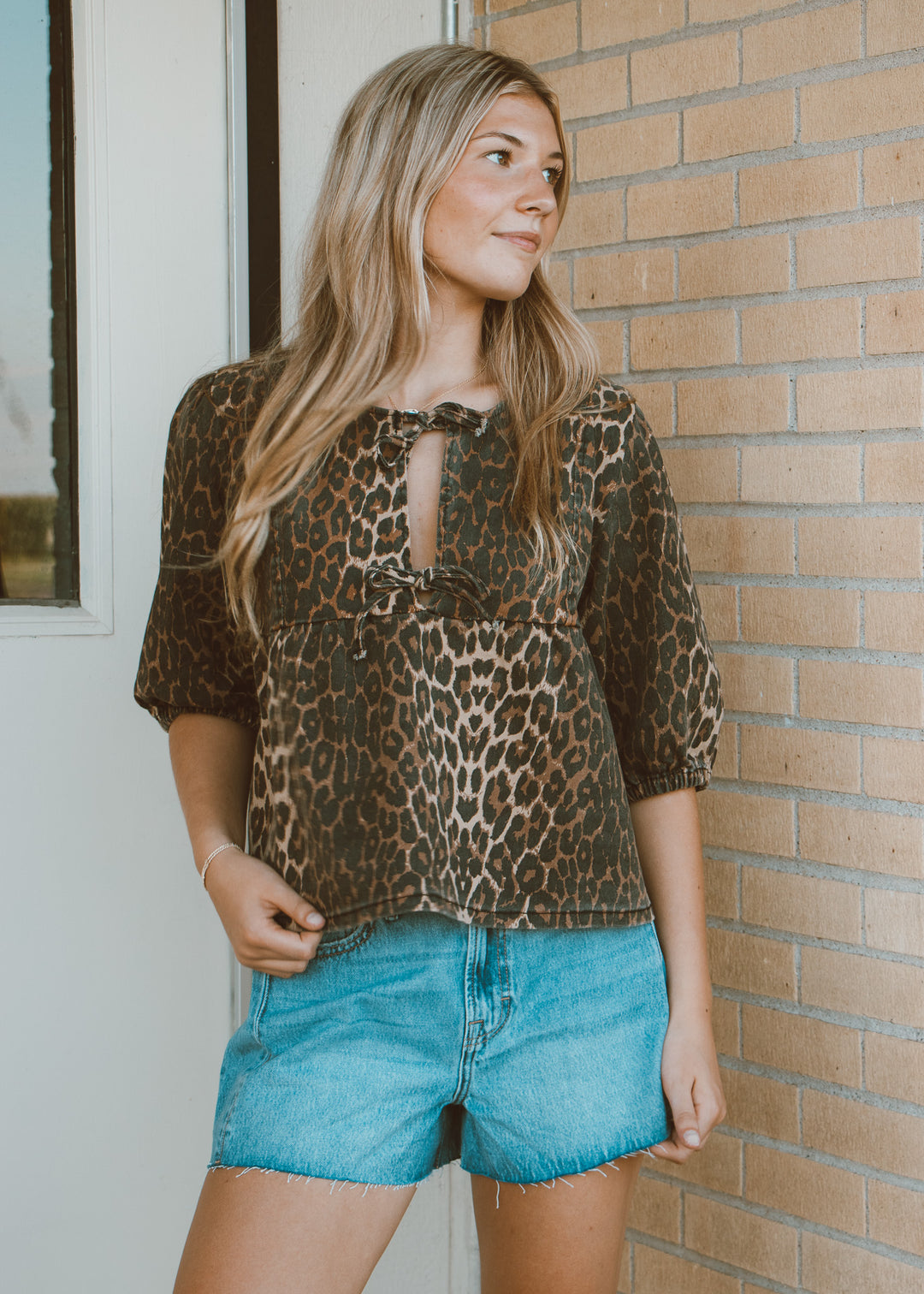Woman wearing a leopard print top and denim shorts standing against a brick wall.