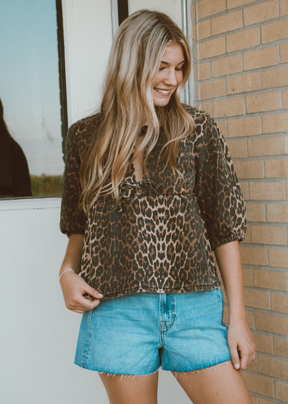 Woman wearing a leopard print top and denim shorts standing against a brick wall.