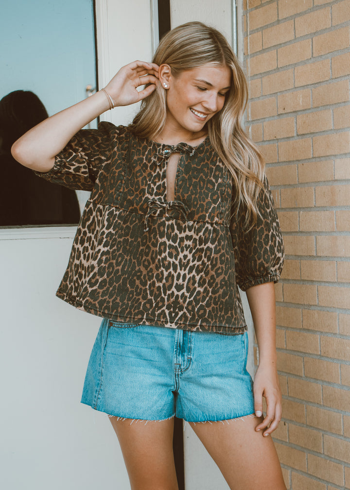 Woman wearing a leopard print top and blue shorts standing against a brick wall.