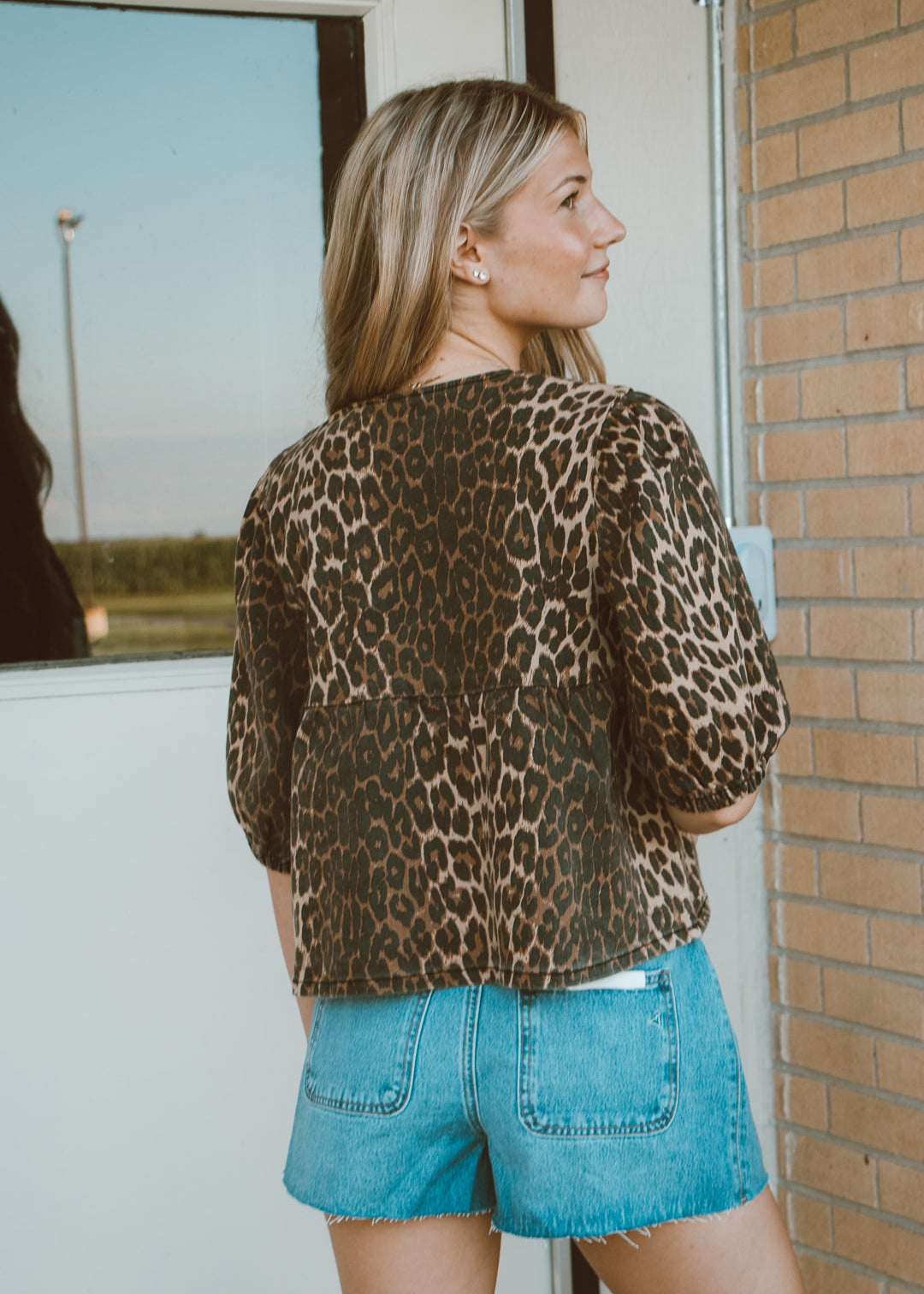 Woman wearing a leopard print jacket and blue shorts standing near a brick wall.