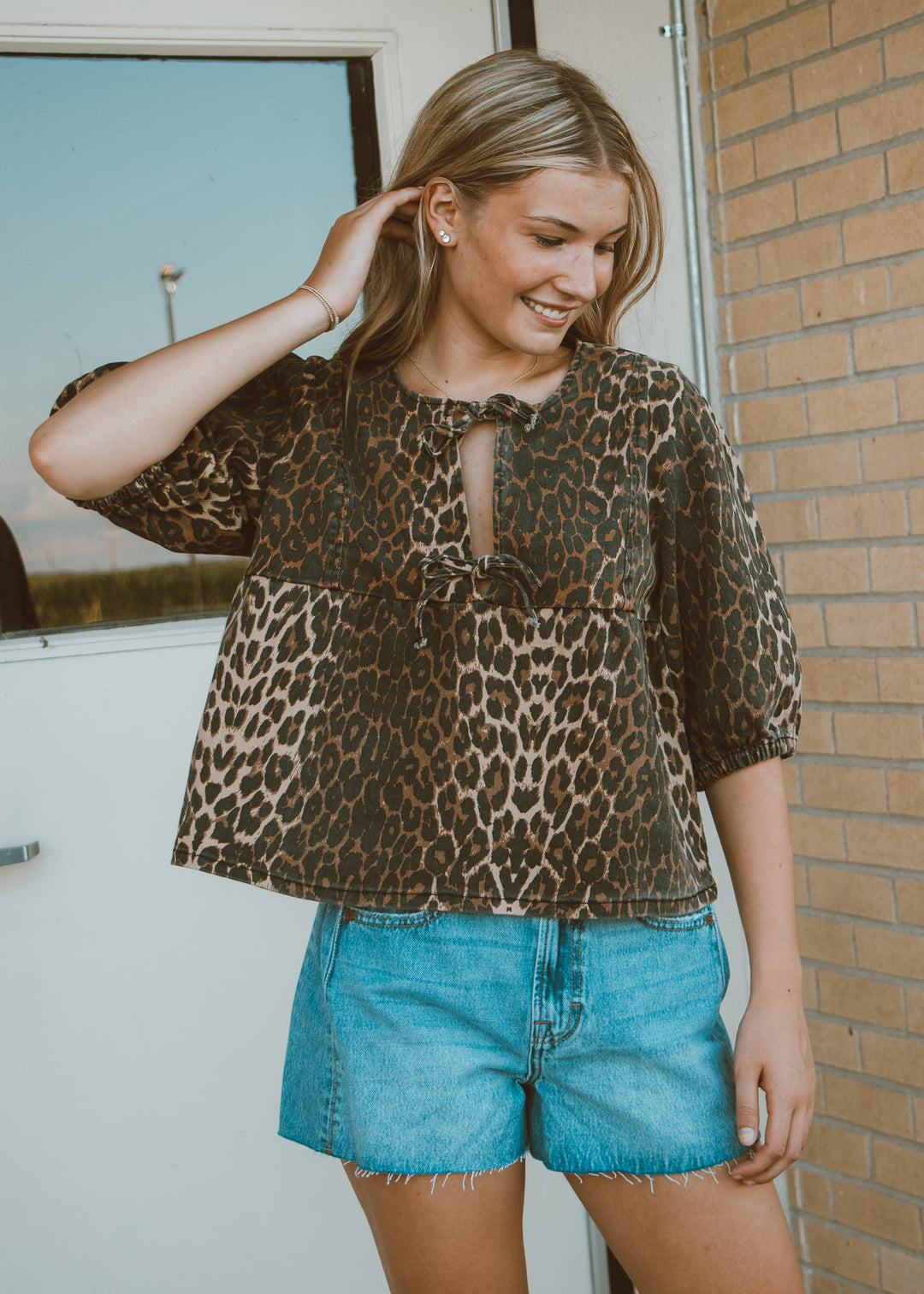 Woman wearing a leopard print top and blue shorts standing against a brick wall.