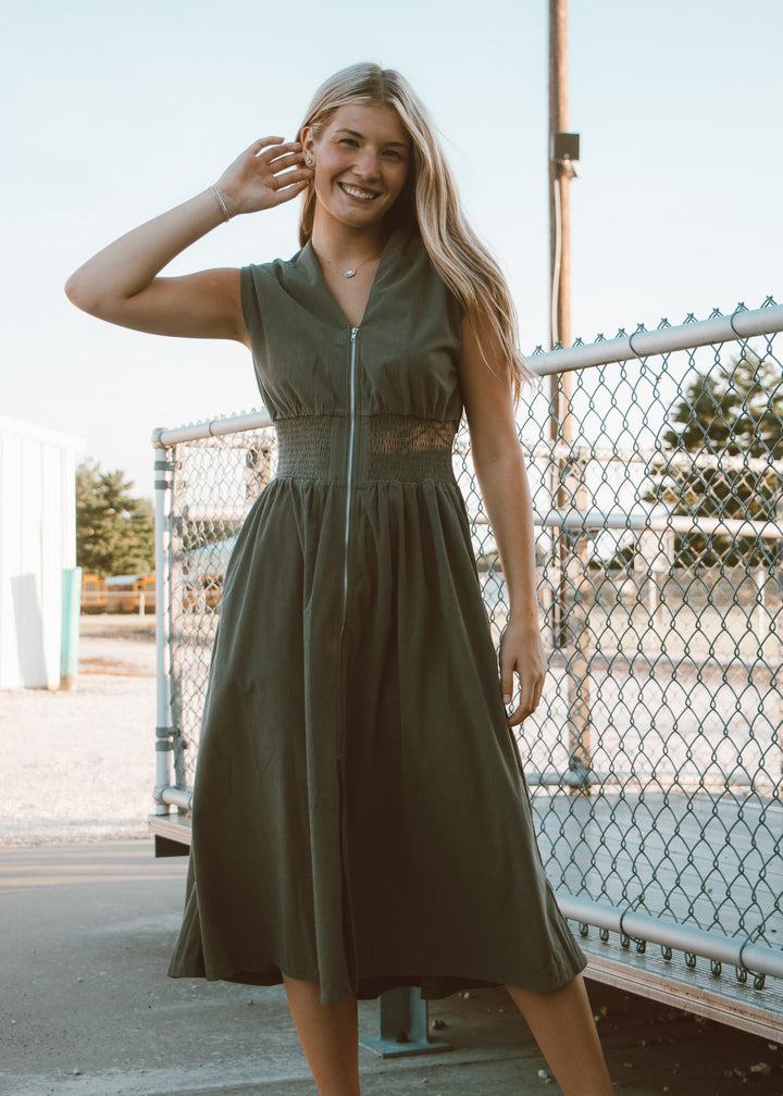 Woman wearing a green dress standing outdoors near a chain-link fence.