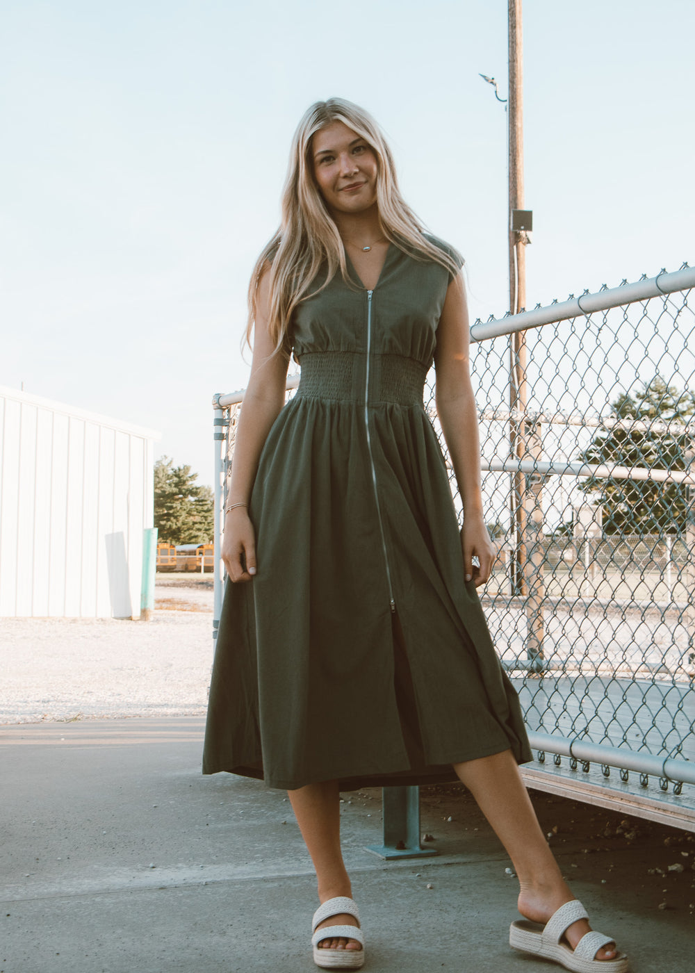 Woman wearing a green dress standing outdoors near a chain-link fence.