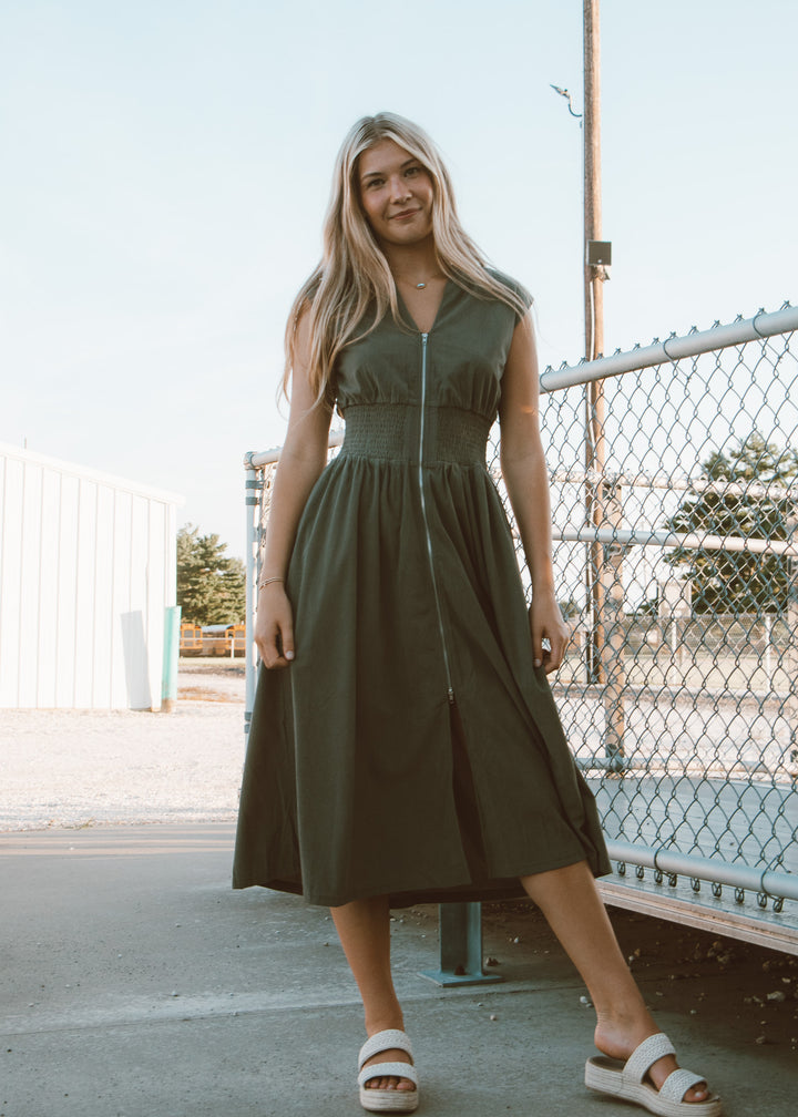 Woman wearing a green dress standing outdoors near a chain-link fence.