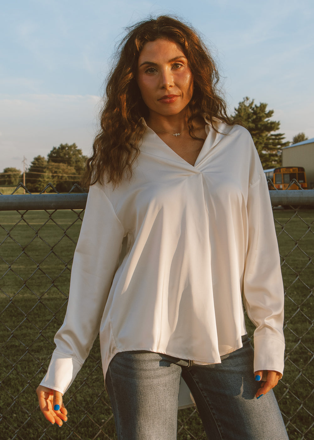 Woman wearing a white blouse and jeans standing in an outdoor setting with a fence and trees in the background.