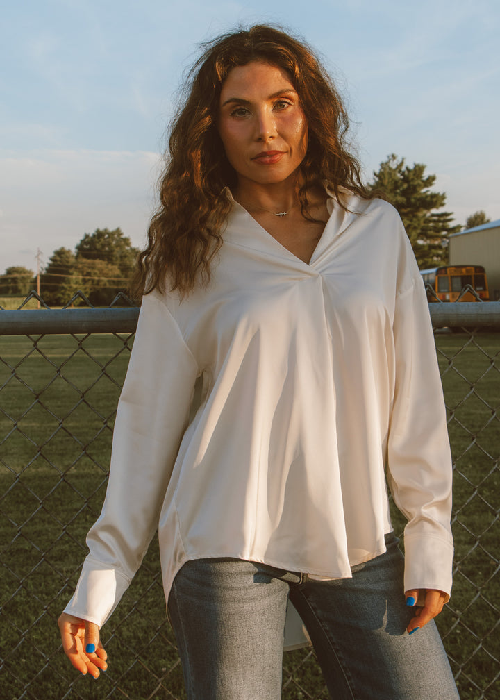 Woman wearing a white blouse and jeans standing in an outdoor setting with a fence and trees in the background.