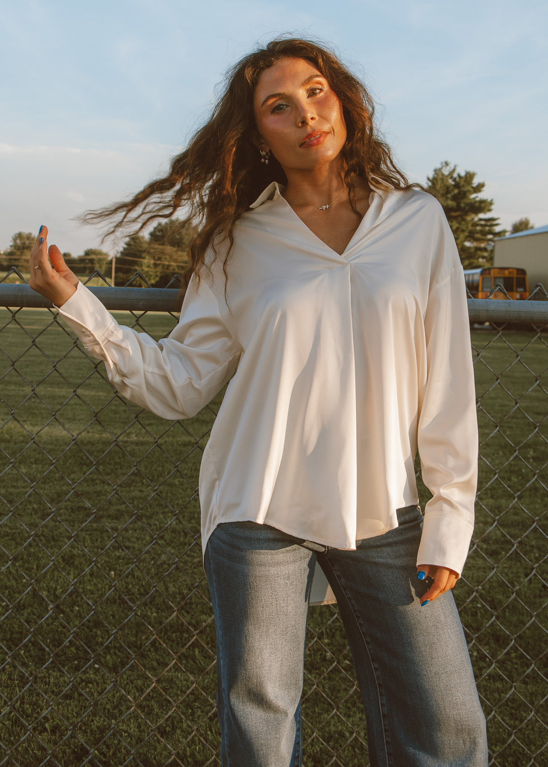 Woman wearing a white blouse and jeans standing outdoors near a fence.