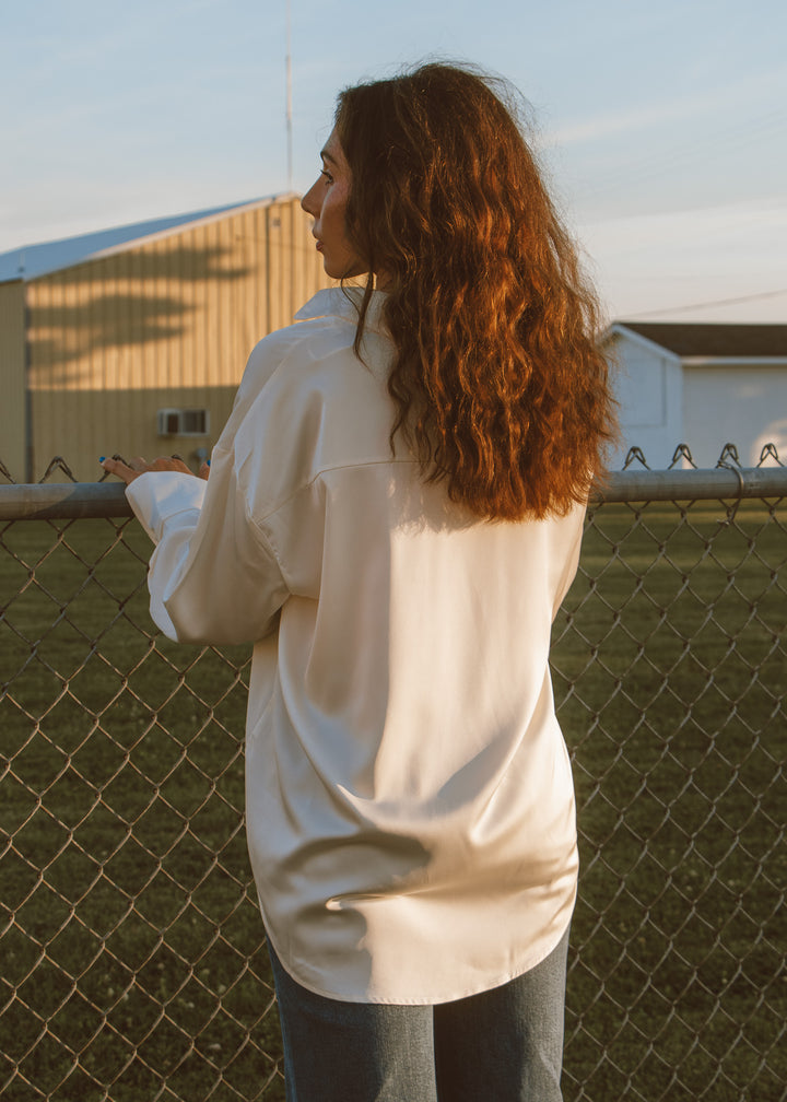 Person wearing a white shirt standing near a chain-link fence with buildings in the background