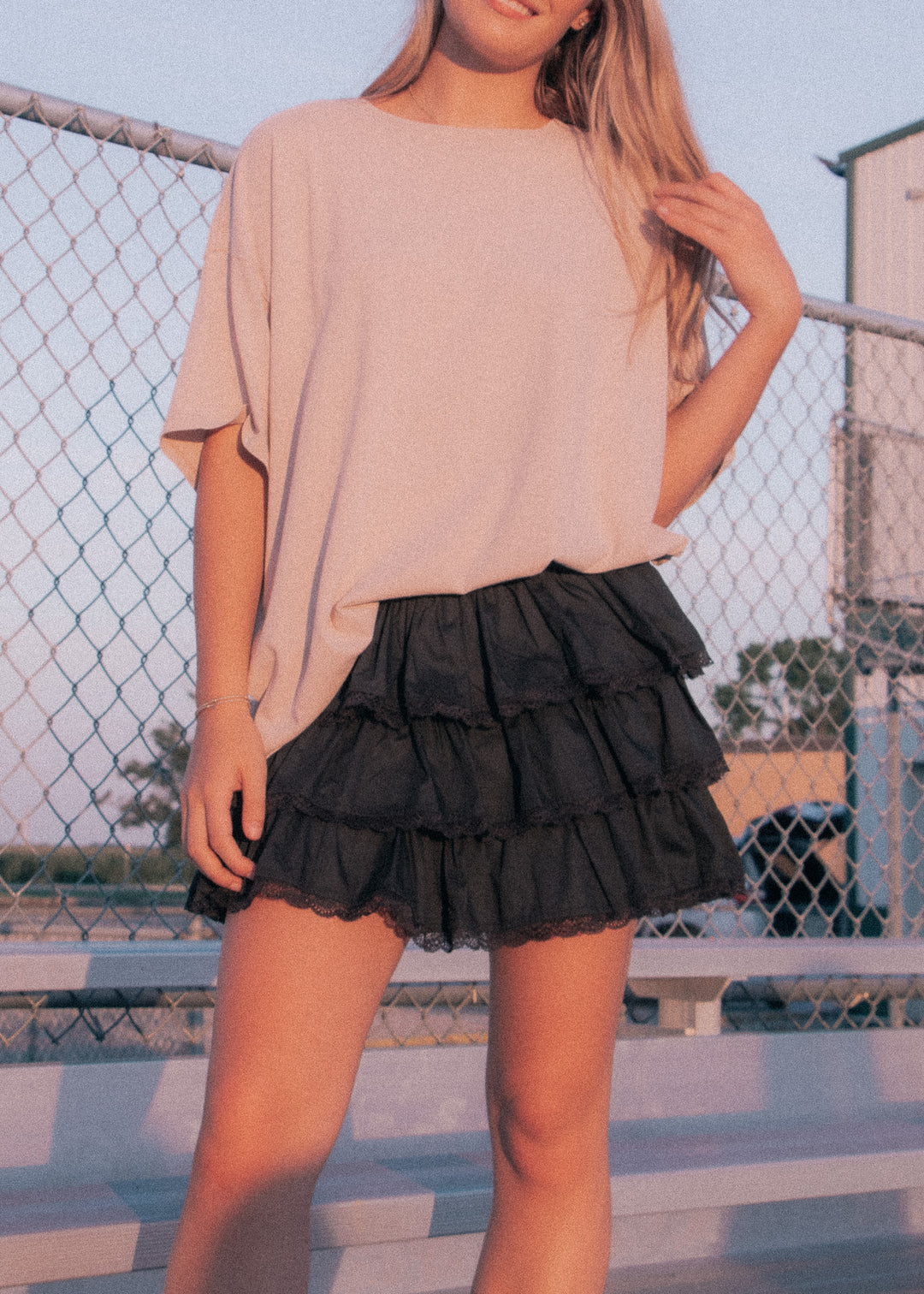 Person wearing a beige top and black tiered skirt standing in front of a chain-link fence.