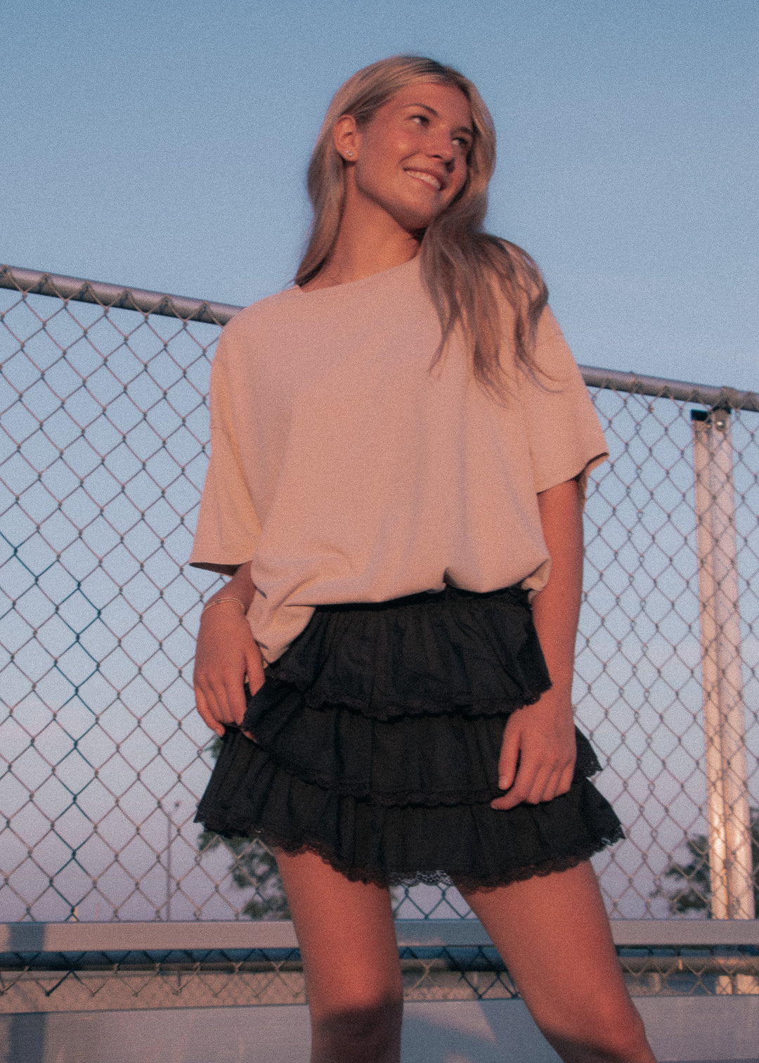 Woman wearing a beige top and black ruffled skirt standing in front of a chain-link fence.