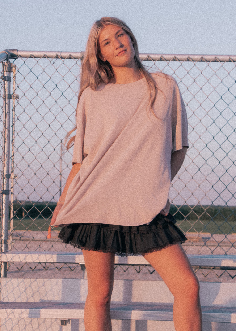 Woman wearing a beige oversized top and black skirt standing in front of a chain-link fence.