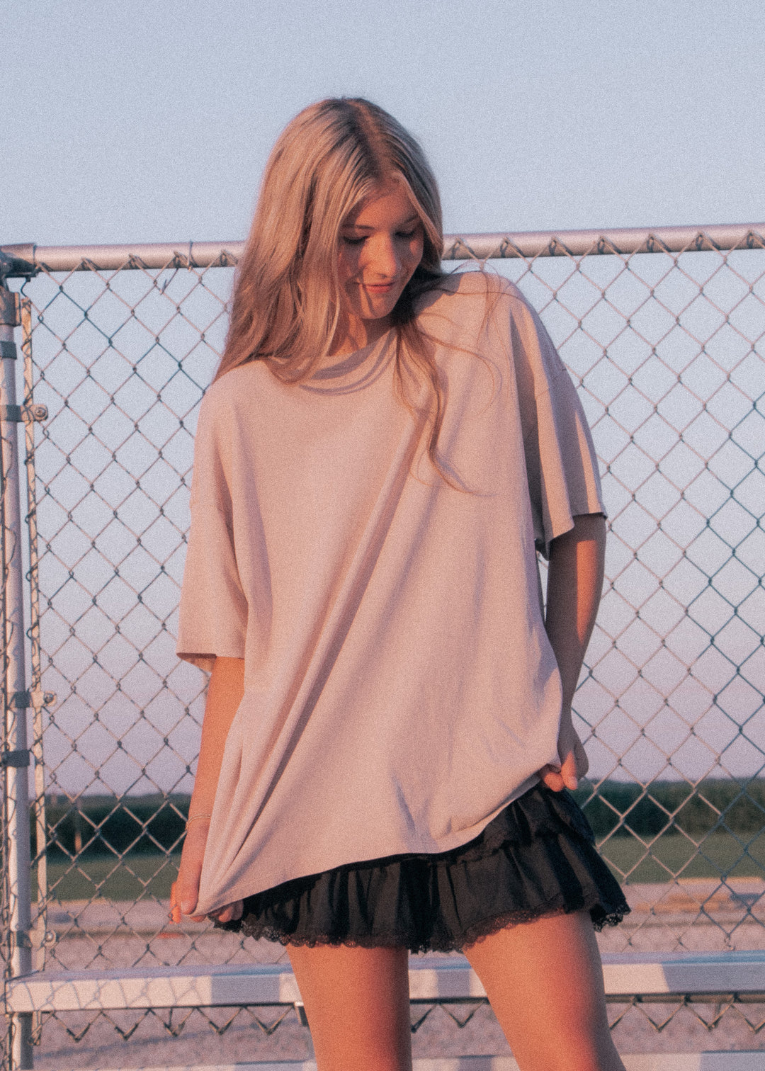 Woman wearing a beige oversized shirt and black skirt standing in front of a chain-link fence.