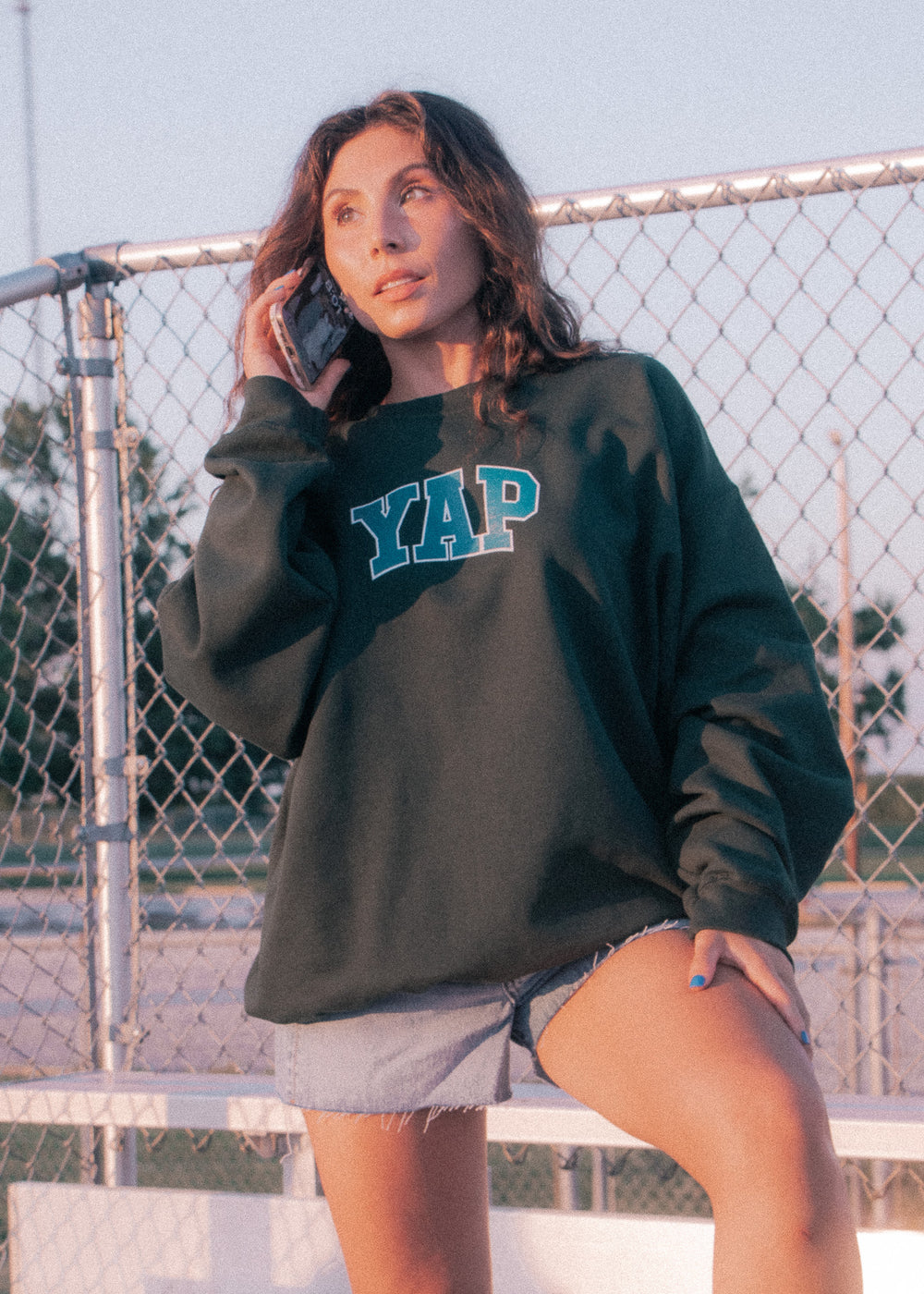 Woman wearing a dark green sweatshirt with 'YAP' text, standing outdoors near a chain-link fence.
