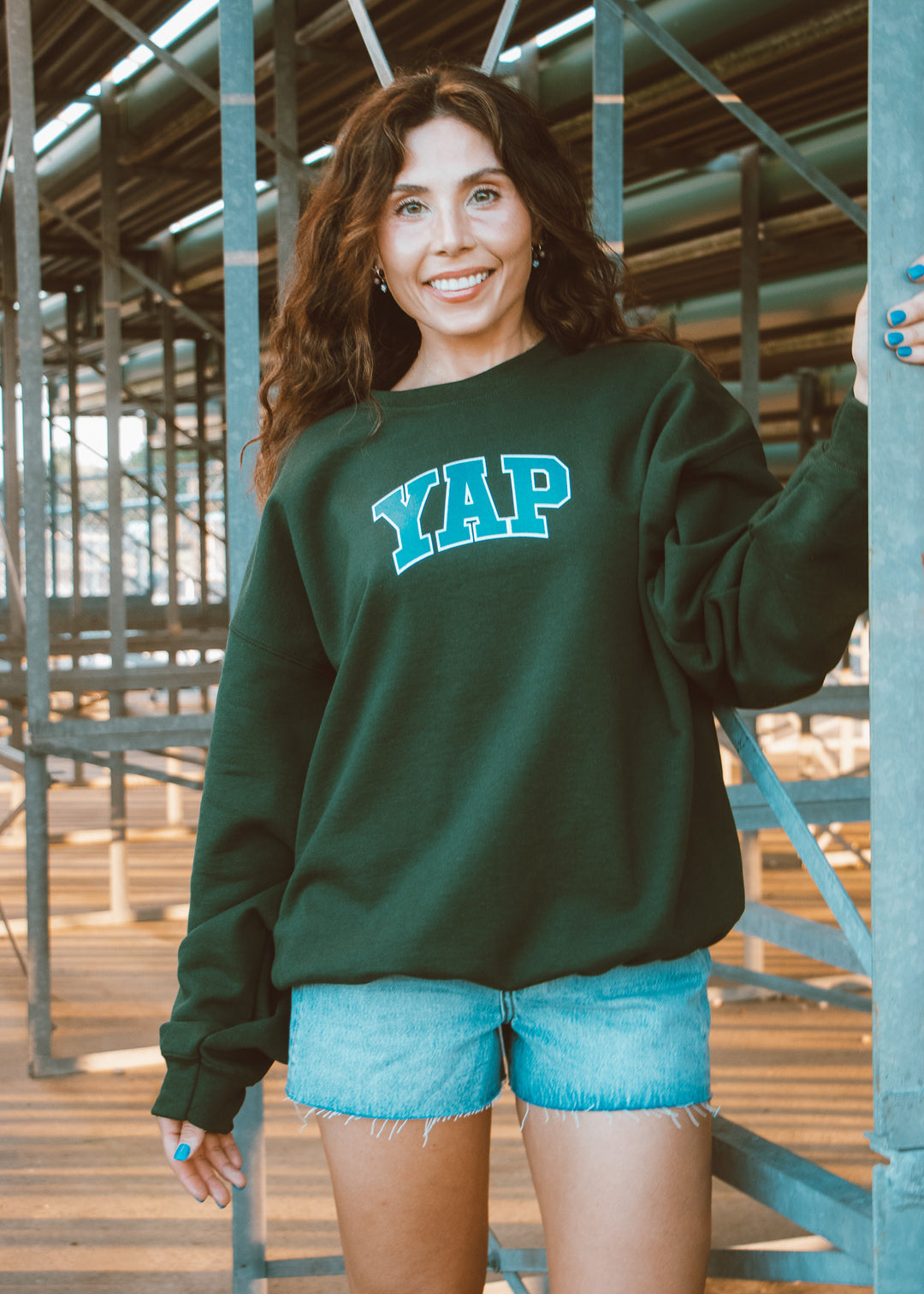 Woman wearing a green sweatshirt with 'YAP' text, standing in an industrial setting.