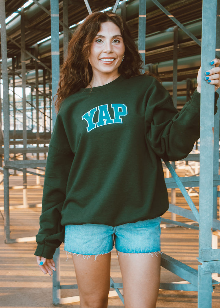Woman wearing a green sweatshirt with 'YAP' text, standing in an industrial setting.