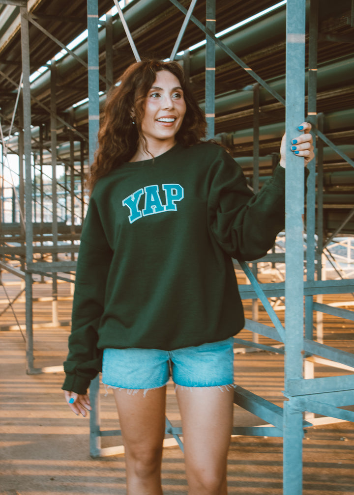 Woman wearing a green sweatshirt with 'YAP' text, standing in an industrial setting.
