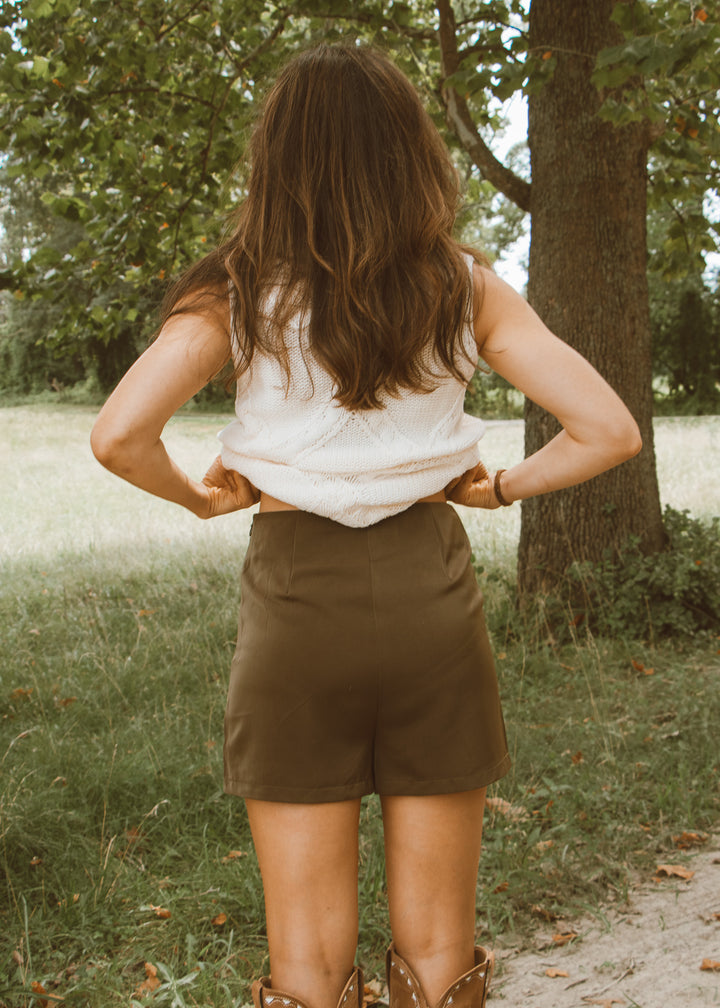 Person wearing a white shirt and brown shorts standing in a natural setting with trees and grass.