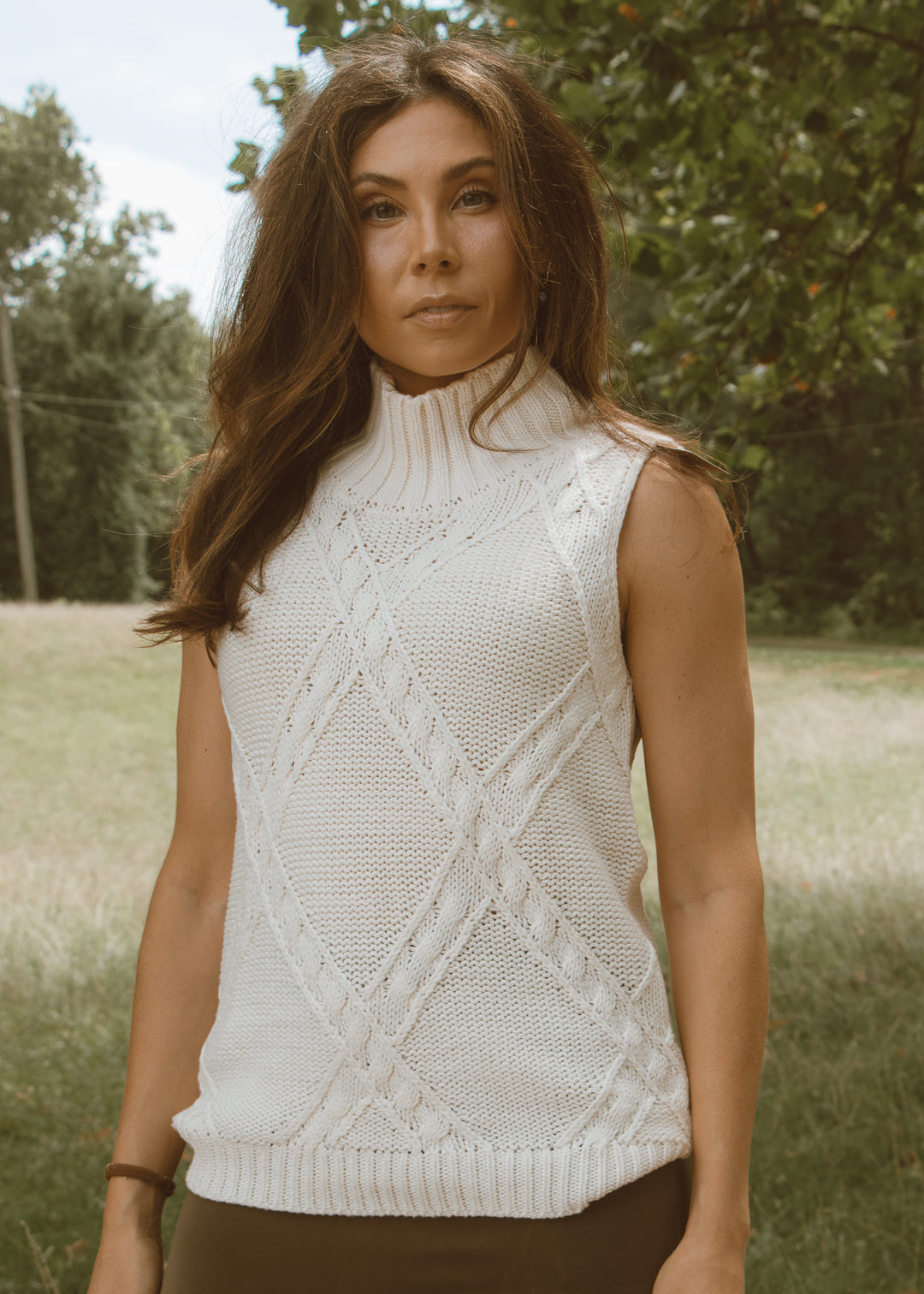 Woman wearing a white sleeveless knitted top outdoors with trees in the background