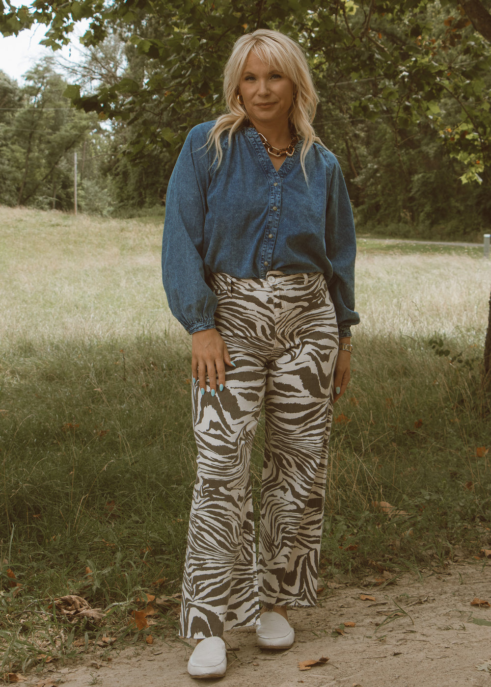Woman wearing a blue shirt and zebra print pants standing in a natural setting with trees and grass.