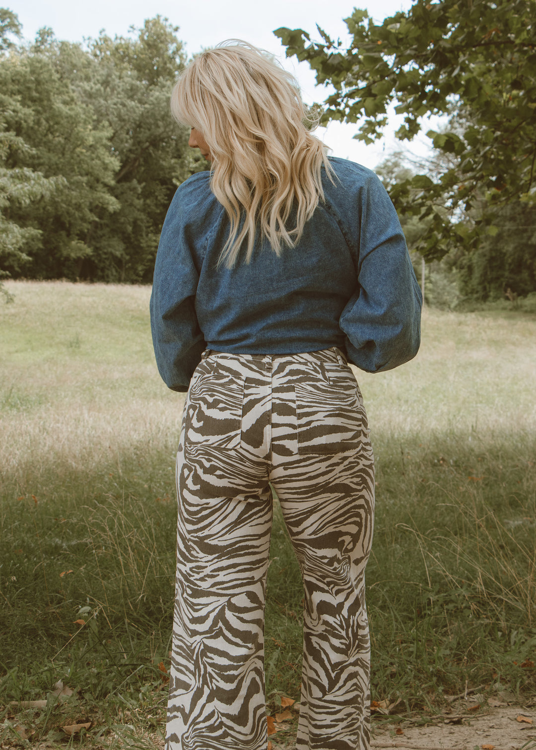 Person wearing a blue top and zebra print pants standing in a grassy field with trees in the background