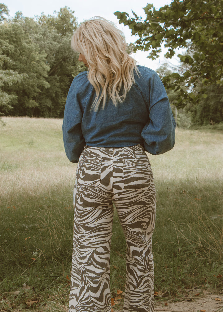 Person wearing a blue top and zebra print pants standing in a grassy field with trees in the background