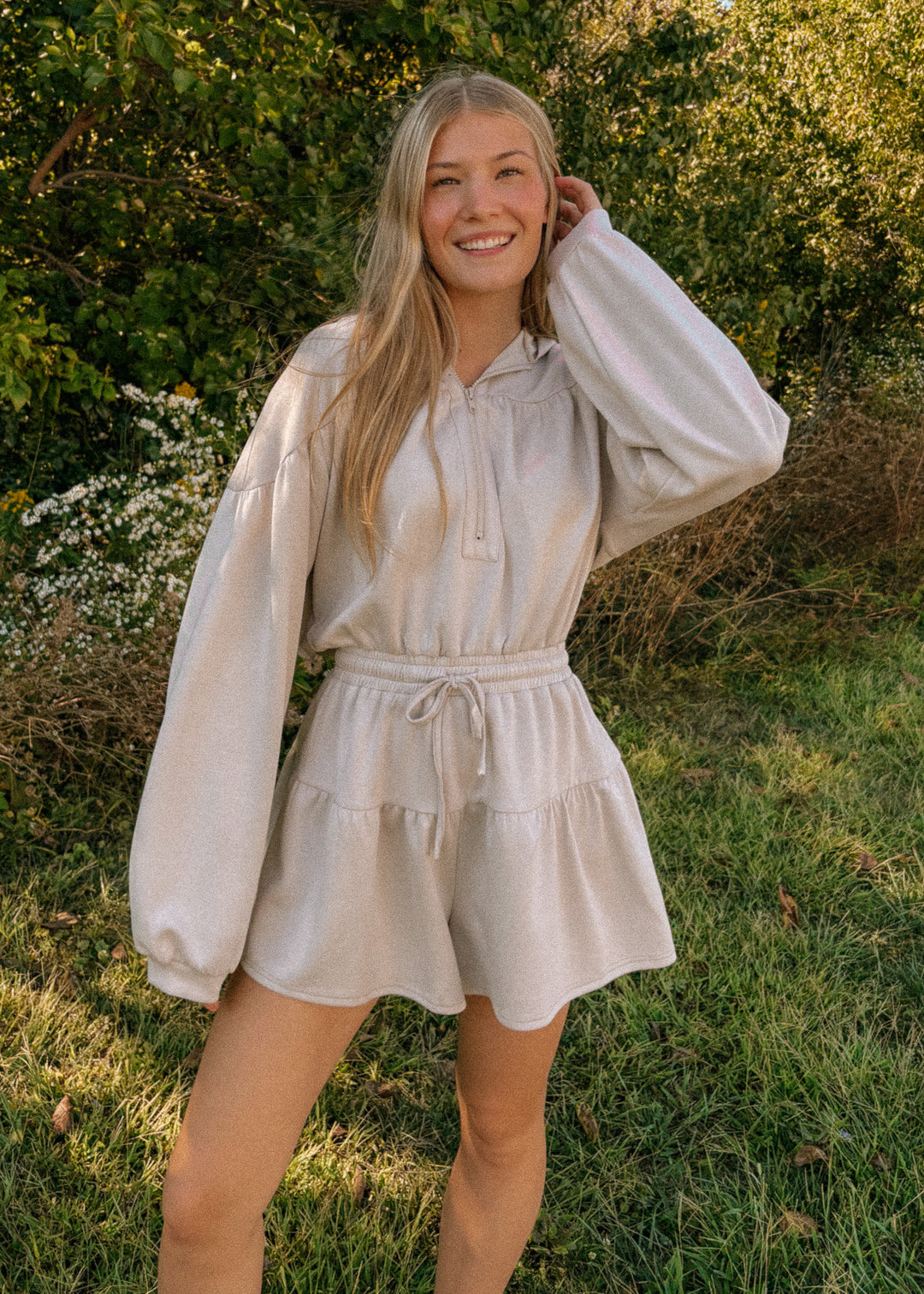 Woman wearing a light-colored romper standing in a natural setting with greenery.