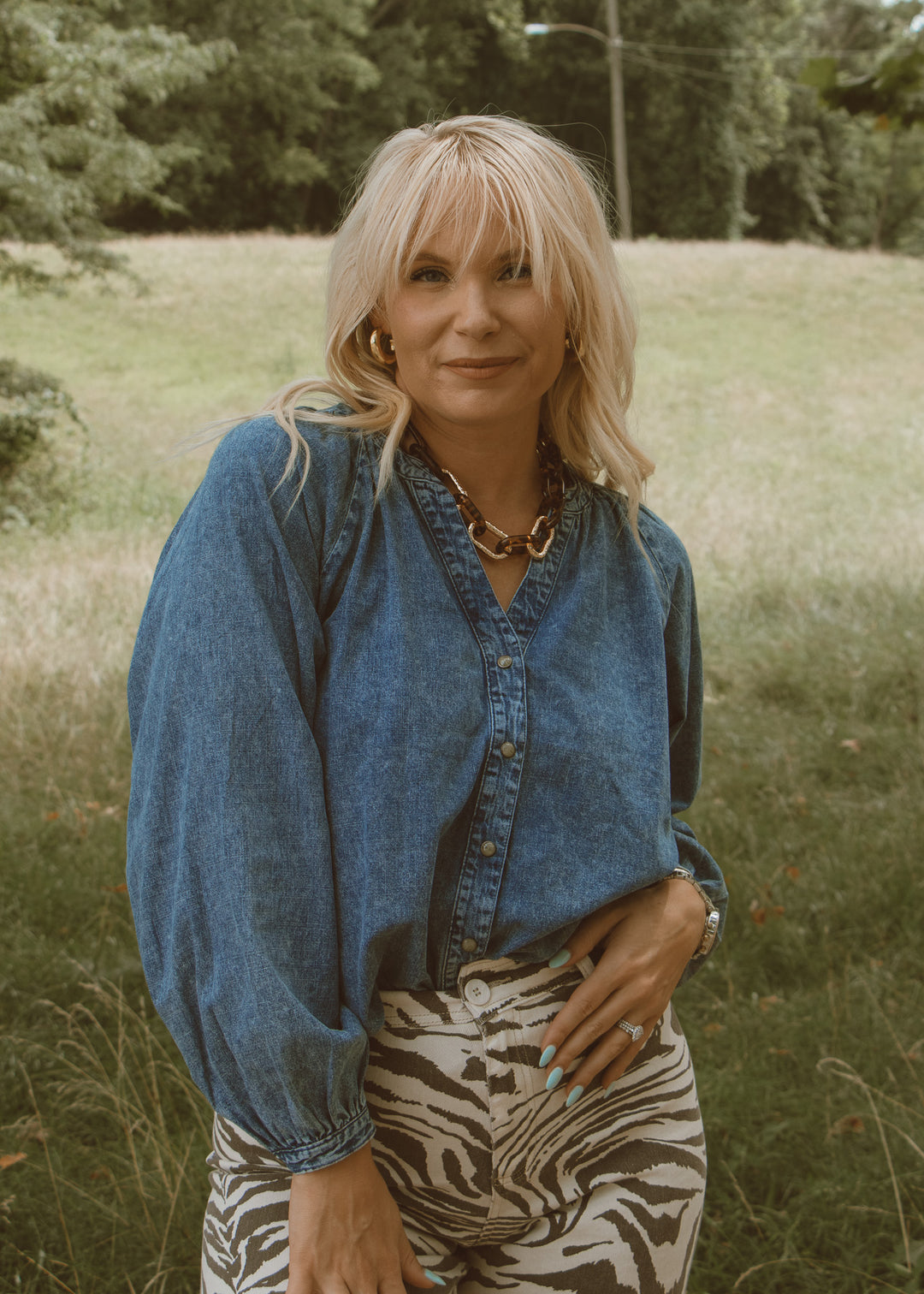 Woman wearing a blue denim shirt and zebra print pants standing in a grassy field.