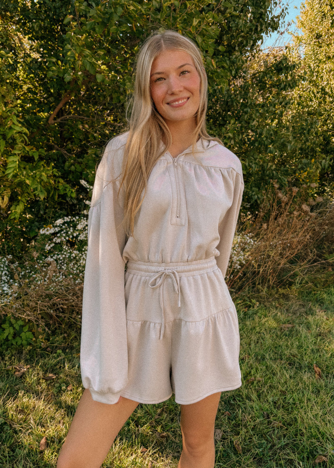 Woman wearing a beige long-sleeve romper standing in a natural setting with trees and grass.