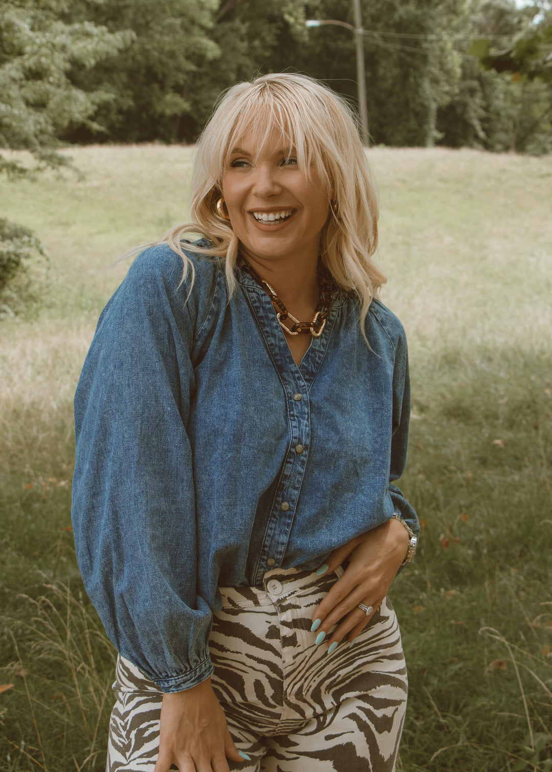 Woman wearing a blue denim shirt and zebra print pants standing in a grassy field.